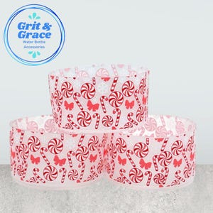 Può includere: Tre accessori per bottiglie d'acqua con un design festivo a bastoncini di zucchero e spirali. Gli accessori sono cilindrici e presentano un motivo ripetuto di bastoncini di zucchero rossi e bianchi, spirali, fiocchi di neve e fiocchi. Il logo Grit & Grace si trova nell'angolo in alto a sinistra.