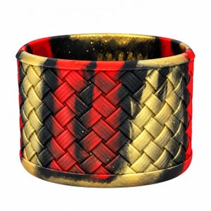 May include: A wide, cylindrical silicone bracelet featuring a textured, woven-look pattern in a marbled blend of red, black, and metallic gold colors. The band has a smooth, solid rim at the top and bottom edges, with a consistent, repeating diagonal weave design.