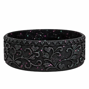 May include: A black silicone ring featuring a raised heart and vine pattern around the exterior. The surface is speckled with small pink and teal dots, and a continuous row of tiny hearts is embossed along the top edge of the band.