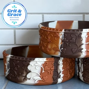 Könnte beinhalten: Ein Set aus runden Silikon-Schutzhüllen für Wasserflaschen mit einem marmorierten braunen, beigen und weißen Farbmuster sowie einem geprägten Herz- und Rankenmuster. Das Logo Grit & Grace Water Bottle Accessories ist oben links zu sehen.