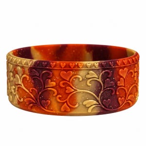 May include: A circular bangle bracelet featuring a marbled blend of orange, red, and tan colors. The exterior surface is embossed with a repeating pattern of swirling vines and small heart shapes, with a row of hearts along the top edge.