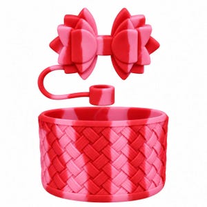 Puede incluir: Un accesorio de silicona para vaso en color rojo y rosa con una textura de tejido de cesta en la base cilíndrica y un lazo decorativo unido por una correa flexible. El artículo presenta un patrón de color jaspeado en todo el material.