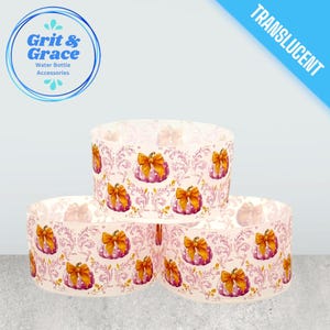 Può includere: Tre accessori traslucidi per bottiglie d'acqua con un motivo ripetuto di zucche rosa con fiocchi arancioni e un design floreale. Gli accessori sono cilindrici e impilati. L'angolo in alto a sinistra presenta un logo con la scritta "Grit & Grace Water Bottle Accessories".