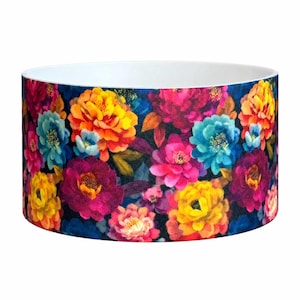 May include: A cylindrical lampshade featuring a vibrant floral pattern against a dark background. The design includes large, detailed blossoms in shades of bright yellow, orange, magenta, and teal, with a clean white interior lining.