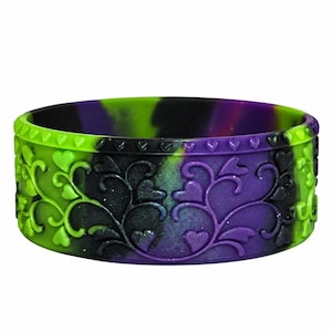 May include: A silicone bracelet featuring a marbled pattern of black, bright green, and purple. The surface is embossed with a repeating vine and heart motif, with a border of small hearts along the top and bottom edges of the band.