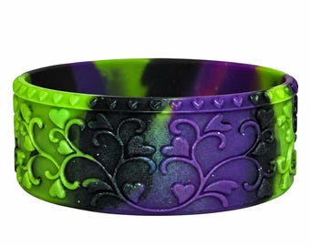 May include: A silicone bracelet featuring a marbled pattern of black, bright green, and purple. The surface is embossed with a repeating vine and heart motif, with a border of small hearts along the top and bottom edges of the band.