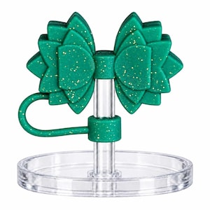 Puede incluir: Un tapón de botella de silicona verde con purpurina que presenta un diseño de lazo en capas en la parte superior. El tapón tiene un poste vertical de plástico transparente y una base circular transparente, con un lazo de silicona verde flexible.