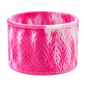 May include: A wide, cylindrical silicone bracelet featuring a vibrant hot pink and white marbled color pattern. The exterior surface is embossed with a repeating vertical leaf or wheat-like texture, framed by a beaded border at the top and bottom.