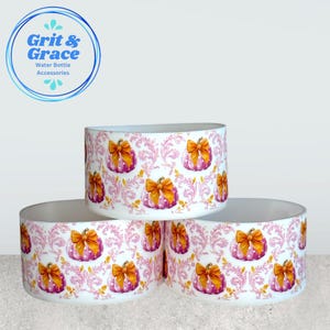 May include: Three cylindrical silicone water bottle boots featuring a repeating pattern of pink pumpkins with white polka dots and orange bows, set against a light pink swirling filigree background. The text Grit & Grace Water Bottle Accessories is visible in the upper left corner.