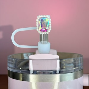 May include: A pink tumbler with a clear lid and a decorative top. The top features a large, iridescent gem surrounded by small, white pearls. A white plastic straw and lid are attached to the tumbler.