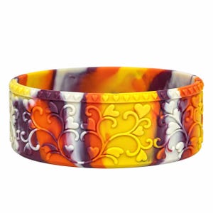 May include: A circular silicone bracelet featuring a marbled pattern of yellow, orange, and deep purple. The exterior surface is embossed with a repeating relief design of swirling vines and small heart shapes along the top edge.