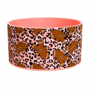 May include: A round pet bowl with a coral pink interior and rim. The exterior features a repeating pattern of brown gingerbread men figures interspersed with a black and pink leopard print design on a light pink background.