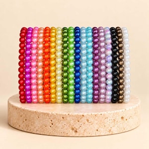 May include: A collection of eighteen individual elastic bracelets arranged in a vertical row, each featuring a single color of smooth, round, glossy beads in a spectrum including red, pink, orange, yellow, green, blue, purple, black, brown, and white.