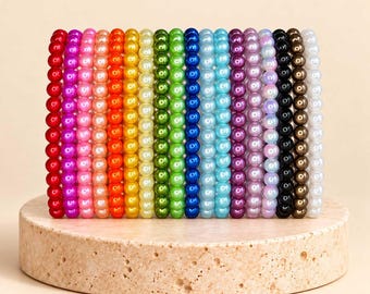 May include: A collection of eighteen individual elastic bracelets arranged in a vertical row, each featuring a single colour of smooth, round, glossy beads in a spectrum including red, pink, orange, yellow, green, blue, purple, black, brown, and white.