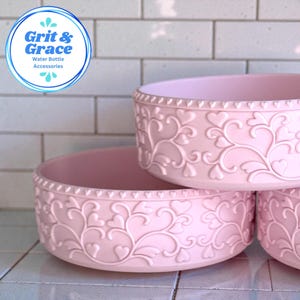Puede incluir: Tres accesorios rosas apilados para botellas de agua. Cada accesorio tiene un patrón de corazones y vides alrededor de los lados. El borde superior tiene una fila de pequeños corazones. El logotipo "Grit & Grace Water Bottle Accessories" está en la esquina superior izquierda.