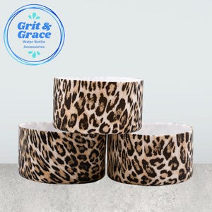 Peut inclure: Trois accessoires cylindriques pour bouteilles d'eau avec un motif léopard. Les accessoires sont de couleur beige, marron et blanc. Le texte "Grit & Grace Water Bottle Accessories" est dans un cercle bleu en haut à gauche.