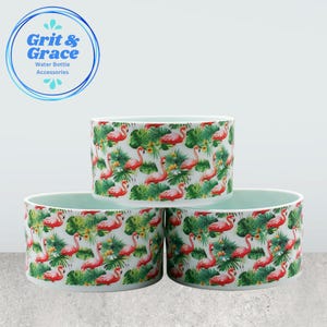 Pode incluir: Três acessórios para garrafas de água com um padrão de flamingos e folhas tropicais. Os acessórios são cilíndricos com um interior azul claro e um exterior branco. O exterior apresenta flamingos cor-de-rosa e folhas verdes sobre um fundo branco. O logótipo "Grit & Grace Water Bottle Accessories" está no canto superior esquerdo.