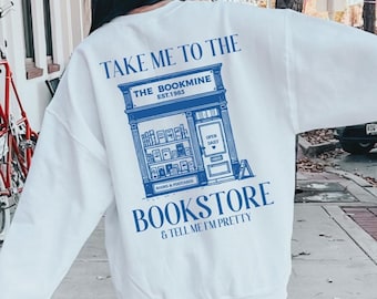 以下が含まれることがあります： 背中に青い本屋のグラフィックが入った白いスウェットシャツ。「TAKE ME TO THE BOOKMINE BOOKSTORE & TELL ME I'M PRETTY」の文字が入っています。スウェットシャツは屋外で着用されています。