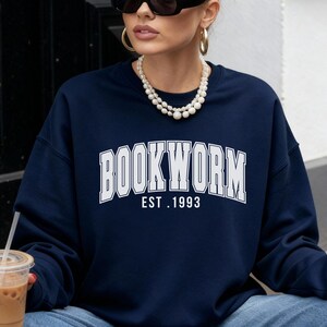 Könnte beinhalten: Ein marineblaues Sweatshirt mit dem Wort "BOOKWORM" in weißen Blockbuchstaben und "EST. 1993" darunter. Die Person trägt eine blaue Jeans und rote Schuhe. Eine Handtasche mit Schlangenmuster und ein Kaffeegetränk sind ebenfalls zu sehen.