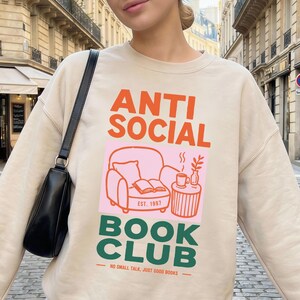 Könnte beinhalten: Ein beiges Langarm-Sweatshirt mit dem Text ANTI SOCIAL BOOK CLUB in Orange und Grün sowie einer Grafik eines Sessels, eines Buches und einer Kaffeetasse. Unter der Grafik steht der Text NO SMALL TALK, JUST GOOD BOOKS.