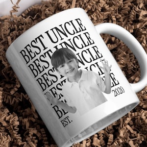 May include: A white ceramic mug featuring a black and white photograph of a child waving. The text BEST UNCLE is repeated vertically in black capital letters, with the year 2020 and the abbreviation EST. printed at the bottom.