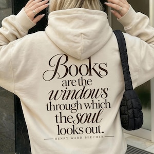 May include: A beige hooded sweatshirt featuring dark brown text on the back that reads: Books are the windows through which the soul looks out. HENRY WARD BEECHER. The sweatshirt is paired with a beige baseball cap and a white pleated skirt.