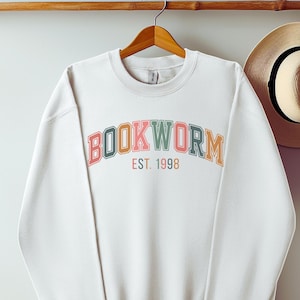 Könnte beinhalten: Weißes Sweatshirt mit Rundhalsausschnitt und dem Wort "BOOKWORM" in bunten, gewölbten Buchstaben. Darunter steht "EST. 1998" in kleinerer Schrift. Das Sweatshirt hängt an einem Holzbügel.