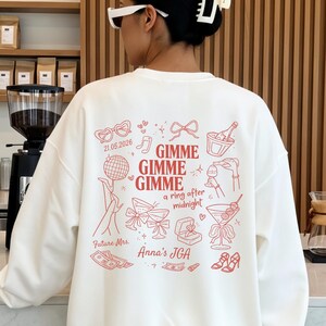May include: A white long-sleeve sweatshirt featuring a red graphic print on the back. The design includes the text "Gimme Gimme Gimme a ring after midnight," "Future Mrs.," "Anna's JGA," and "21.05.2026," surrounded by illustrations of a disco ball, rings, and drinks.