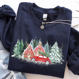 May include: A navy blue crewneck sweatshirt featuring a graphic print of a red vintage car parked in front of two red houses surrounded by snow-covered evergreen trees. The sweatshirt is paired with light blue distressed denim jeans.