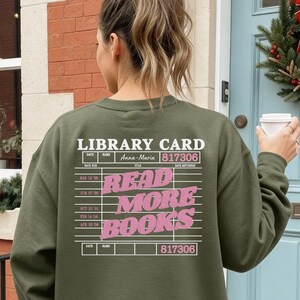 Könnte beinhalten: Ein olivgrünes Sweatshirt mit Rundhalsausschnitt und einer weiß-rosa Grafik auf der Rückseite. Das Design imitiert eine alte Bibliothekskarte mit dem Text "LIBRARY CARD", "Anna-Maria", "817306" und dem fettgedruckten rosa Schriftzug "READ MORE BOOKS".