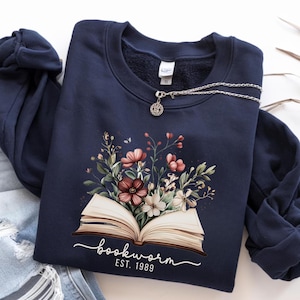 May include: A navy blue sweatshirt with a floral design above an open book, with the text "bookworm est. 1989" underneath. A silver necklace with a pendant rests on the sweatshirt, which is placed on blue jeans.