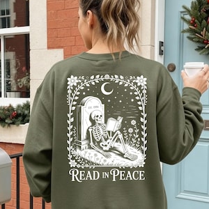 Könnte beinhalten: Ein olivgrünes Sweatshirt mit Rundhalsausschnitt und einem weißen Grafikdruck auf der Rückseite. Das Motiv zeigt ein Skelett, das an einem Grabstein mit der Aufschrift EST. 1996 lehnt und ein Buch unter einem Halbmond liest, mit dem Text READ IN PEACE.