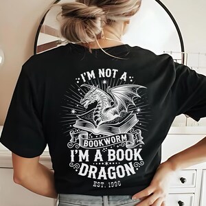 Könnte beinhalten: Schwarzes T-Shirt mit einem weißen Drachen-Motiv auf einem Buch und dem Text "I'M NOT A BOOKWORM I'M A BOOK DRAGON EST. 1996". Das Design zeigt einen Drachen mit Flügeln und Sternen.