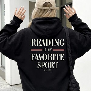 Könnte beinhalten: Ein schwarzer Kapuzenpullover mit weißem Text auf der Rückseite, der READING IS MY FAVORITE SPORT lautet, mit dem Text EST. 1995 darunter. Der Hoodie wird mit einer beigen Baseballkappe, einem weißen Faltenrock und einer schwarzen Stepptasche getragen.