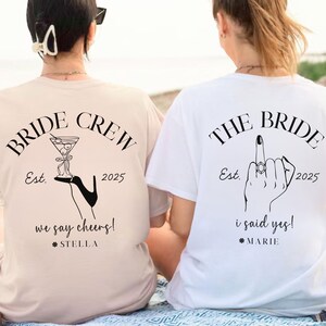 May include: Two women wearing matching t-shirts with wedding-themed graphics. One shirt is beige with "BRIDE CREW" and a cocktail illustration. The other is white with "THE BRIDE" and a hand gesture. Both shirts have the year 2025.