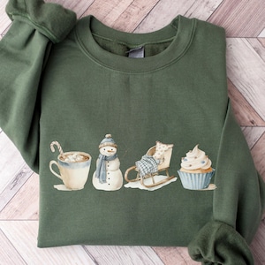 Könnte beinhalten: Ein olivgrünes Sweatshirt mit Rundhalsausschnitt und einem Grafikdruck, der eine Tasse mit Zuckerstange, einen Schneemann mit blauem Schal und Mütze, einen Holzschlitten mit karierter Decke sowie einen Cupcake mit blauem Zuckerguss zeigt.