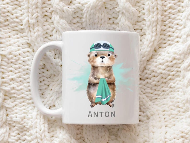 Pode incluir: Caneca de cer&acirc;mica branca com uma ilustra&ccedil;&atilde;o em aquarela de uma lontra usando touca e &oacute;culos de nata&ccedil;&atilde;o, segurando uma toalha verde-azulado. O nome "ANTON" &eacute; impresso em preto na parte inferior da caneca. A caneca tem uma al&ccedil;a branca.