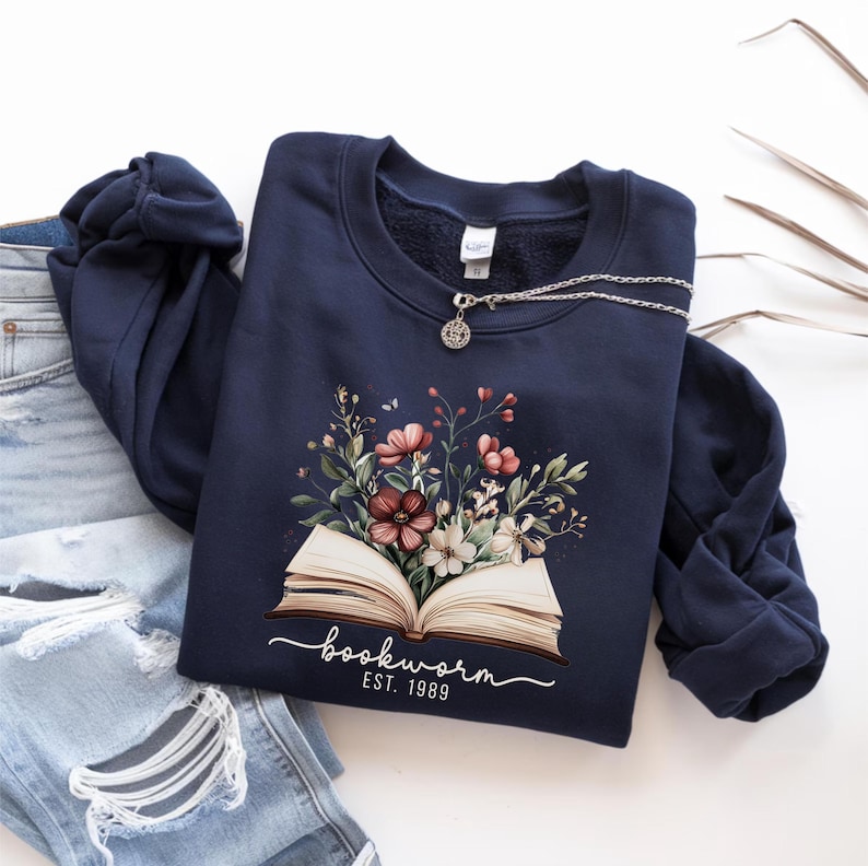 May include: A navy blue sweatshirt with a floral design above an open book, with the text "bookworm est. 1989" underneath. A silver necklace with a pendant rests on the sweatshirt, which is placed on blue jeans.