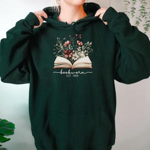 May include: Dark green hoodie with a floral and book design. The design features the word "bookworm" in script, with "EST. 1989" below. The hoodie has a front pocket and a drawstring hood. A casual garment.