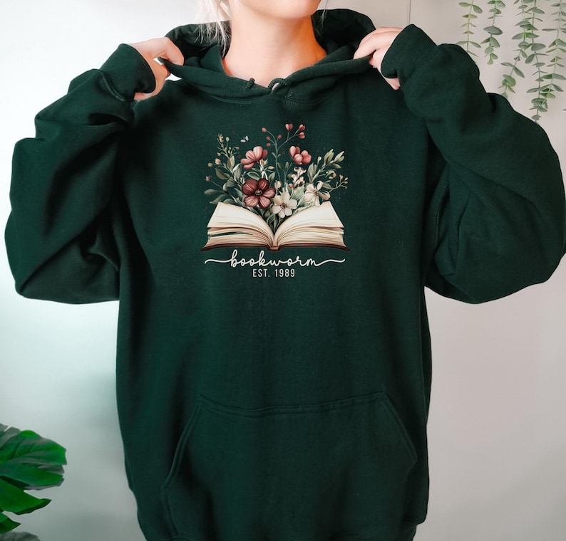 May include: Dark green hoodie with a floral and book design. The design features the word "bookworm" in script, with "EST. 1989" below. The hoodie has a front pocket and a drawstring hood. A casual garment.