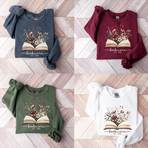 May include: Four sweatshirts in navy, burgundy, olive green, and white. Each has a floral design sprouting from an open book, with the word "bookworm" and "EST. 1989" printed below. A cozy, casual style.