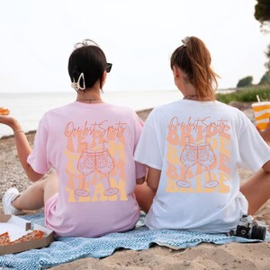 May include: Two oversized t-shirts, one pink and one white, with the text "One last Spritz" and a graphic of two cocktails. The shirts are worn by people sitting on a beach, suggesting a vacation or party theme.
