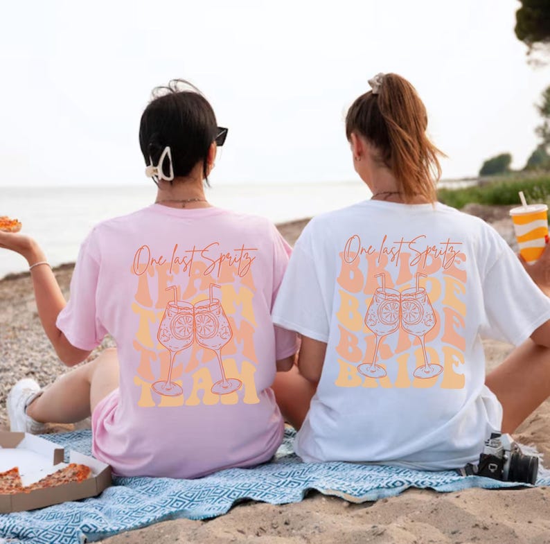 May include: Two oversized t-shirts, one pink and one white, with the text "One last Spritz" and a graphic of two cocktails. The shirts are worn by people sitting on a beach, suggesting a vacation or party theme.