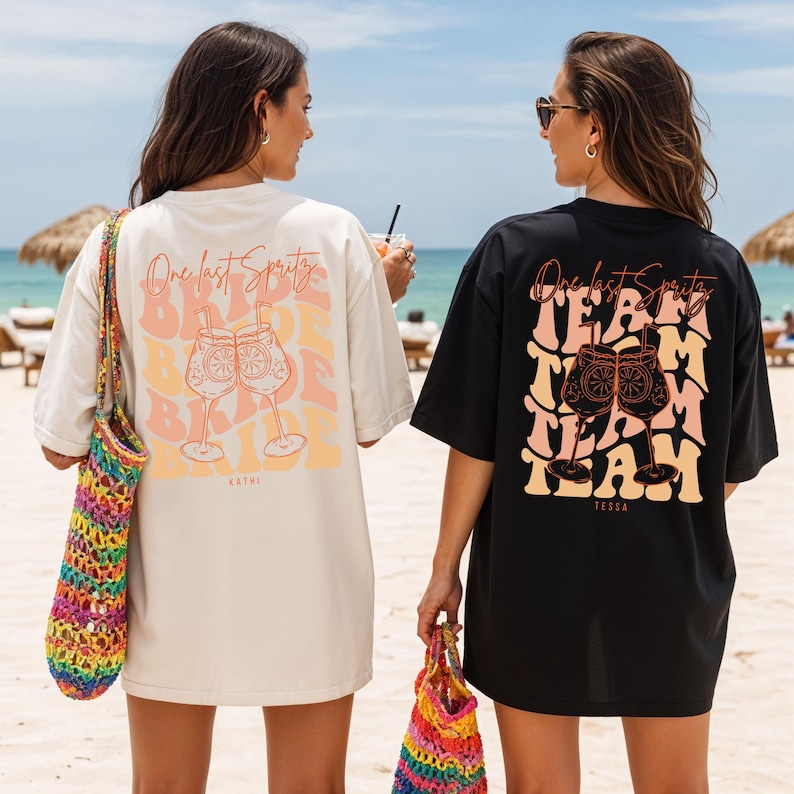 May include: Two oversized t-shirts, one cream and one black, with a graphic design on the back. The design features the text "One Last Spritz" and "Bride Babe" or "Team Team" with cocktail glasses. The shirts are worn on a beach.