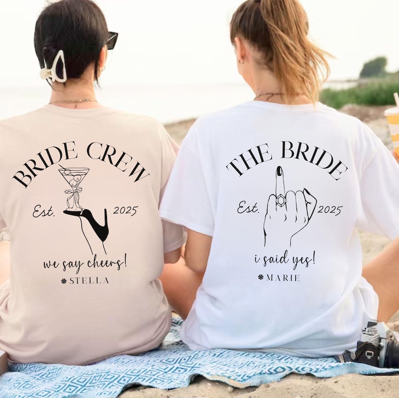 May include: Two women wearing matching t-shirts with wedding-themed graphics. One shirt is beige with "BRIDE CREW" and a cocktail illustration. The other is white with "THE BRIDE" and a hand gesture. Both shirts have the year 2025.