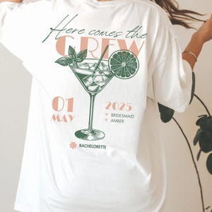 May include: White t-shirt with a green and pink graphic of a cocktail glass with a lime wedge and the text "Here comes the CREW 01 MAY 2025 *BRIDESMAID *AMBER *BACHELORETTE"