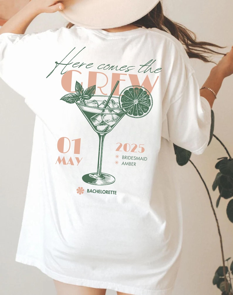 May include: White t-shirt with a green and pink graphic of a cocktail glass with a lime wedge and the text "Here comes the CREW 01 MAY 2025 *BRIDESMAID *AMBER *BACHELORETTE"