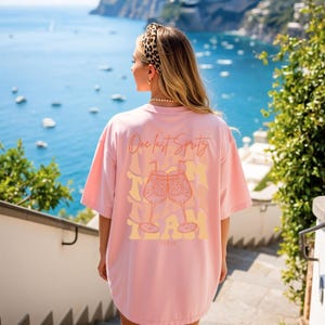 May include: A light pink t-shirt with the text "One last Spritz" and a graphic of two cocktail glasses. The word "TEAM" is printed below the glasses. The shirt is oversized and has short sleeves.