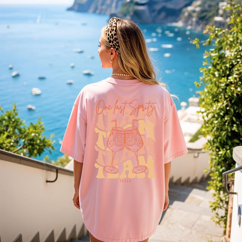 May include: A light pink t-shirt with the text "One last Spritz" and a graphic of two cocktail glasses. The word "TEAM" is printed below the glasses. The shirt is oversized and has short sleeves.