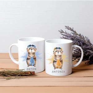 Pode incluir: Duas canecas de cer&acirc;mica branca com ilustra&ccedil;&otilde;es de lontras. Cada lontra usa &oacute;culos de nata&ccedil;&atilde;o e segura uma toalha. Uma caneca diz "ANTON" e a outra "ANTONIA". As lontras est&atilde;o sobre um fundo de aquarela azul e amarelo.